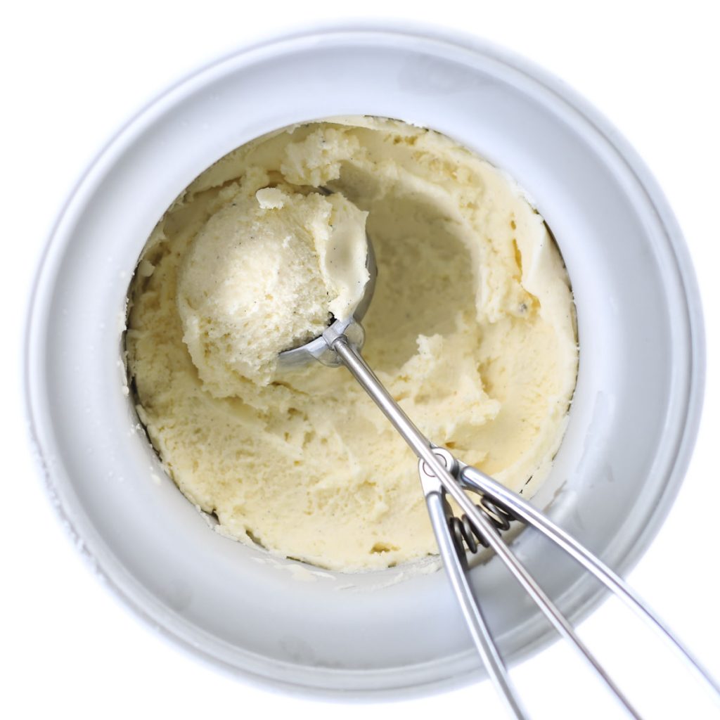 A scoop filled with vanilla bean ice cream placed in an ice cream maker bowl with vanilla bean ice cream, seen from above