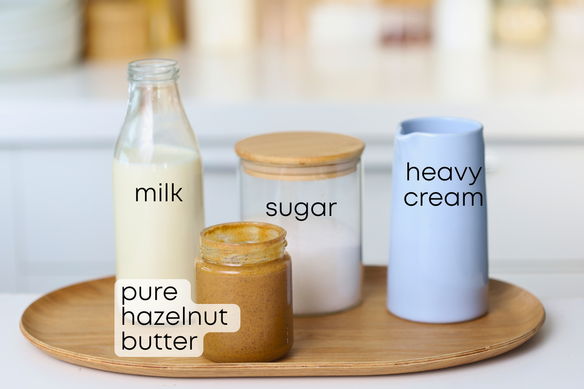Four ingredients for ultra-creamy hazelnut ice cream arranged on a wooden tray: a glass bottle of milk, a jar of sugar, a jug of heavy cream, and a jar of pure hazelnut butter. Each item is clearly labeled.
