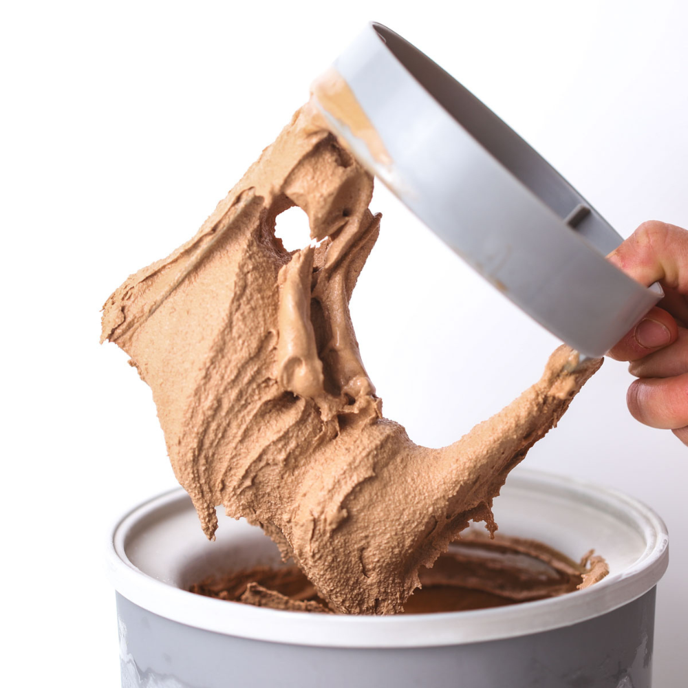 Chocolate-only ice cream made with xanthan gum being lifted from a tub with a spatula, stretching to show its creamy texture.